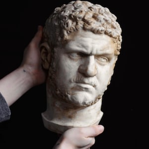 May include: A detailed bust of a man with short, curly hair and a stern expression. The sculpture is off-white with subtle brown shading, suggesting an aged appearance. The bust is held against a black background.