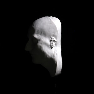 May include: A white plaster cast of a person's head, showing the profile of the face. The cast is detailed and shows the features of the face, including the nose, mouth, and ears.