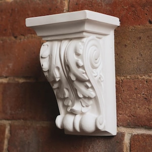 May include: A white, decorative corbel featuring intricate leaf and scroll patterns. The architectural bracket is mounted on a red brick wall, showcasing its classical design. The corbel is made of plaster.