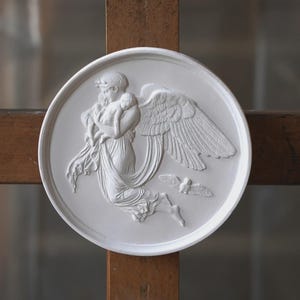 May include: A white circular relief sculpture depicting a classical angel figure holding a baby, with detailed wings and a flowing gown. Two small birds are shown in flight. The sculpture is set against a wooden background.