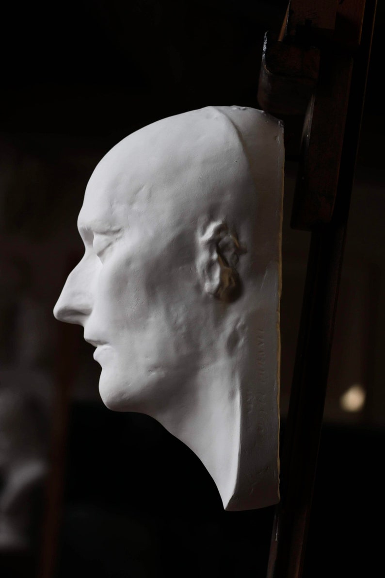 May include: A white plaster cast of a person's face, viewed from the side. The cast is mounted on a wooden easel.