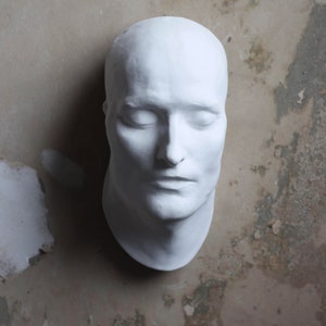 May include: A white plaster cast of a human face with closed eyes. The cast is on a textured, grey background.