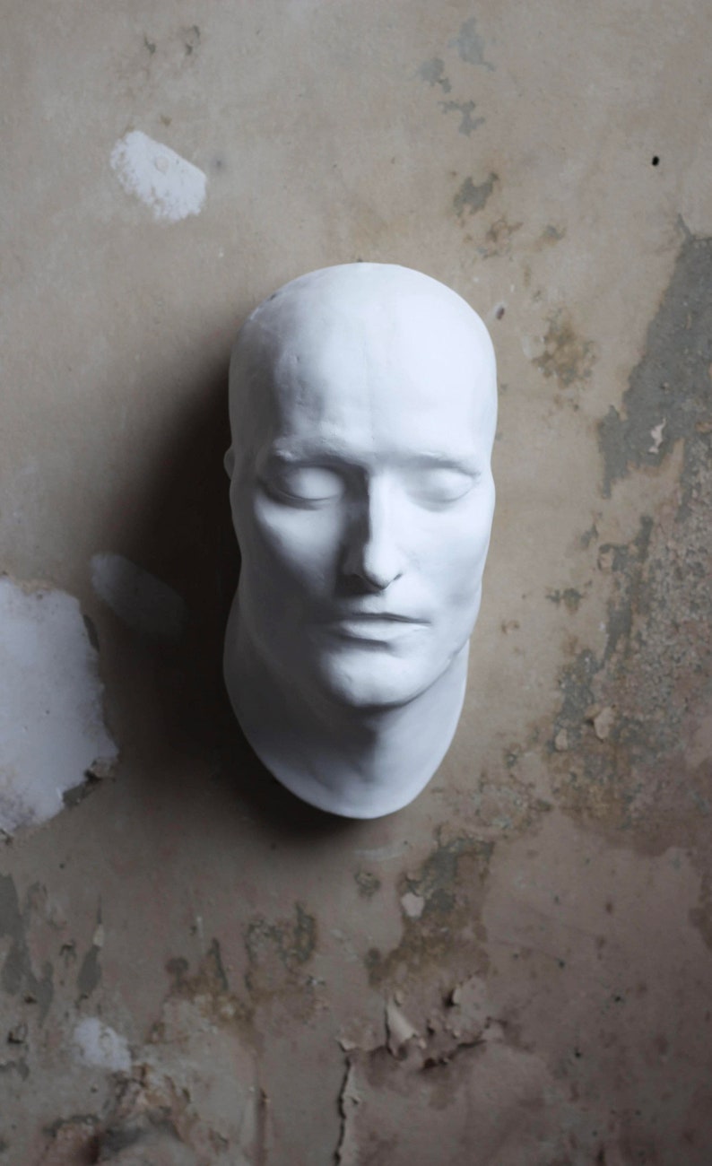 May include: A white plaster cast of a human face with closed eyes. The cast is on a textured, grey background.