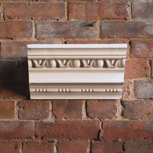 May include: A section of decorative crown molding, painted in cream and brown tones, featuring a repeating pattern of stylized leaves and spheres. The molding is set against a rustic red brick wall, showcasing its architectural detail.