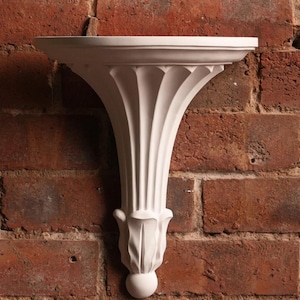 May include: A white, ornate wall shelf with a decorative, fluted support and a round base, mounted on a red brick wall. The shelf has a classic, architectural design, perfect for displaying small items. The shelf's dimensions are not visible.