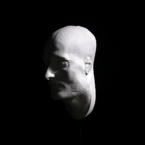May include: A white plaster cast of a person's head, showing the profile of the face. The cast is mounted on a black stand.