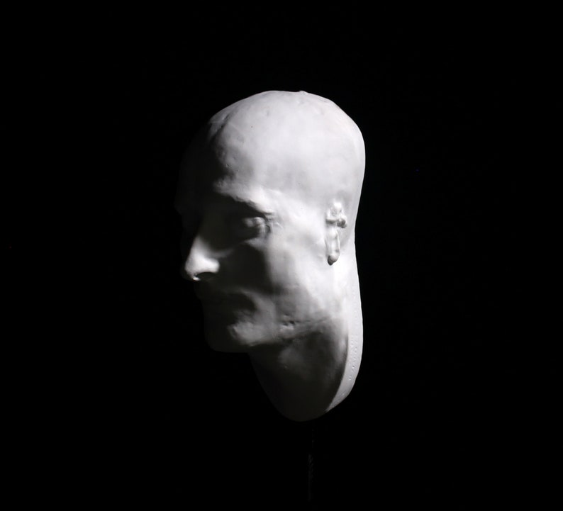 May include: A white plaster cast of a person's head, showing the profile of the face. The cast is mounted on a black stand.