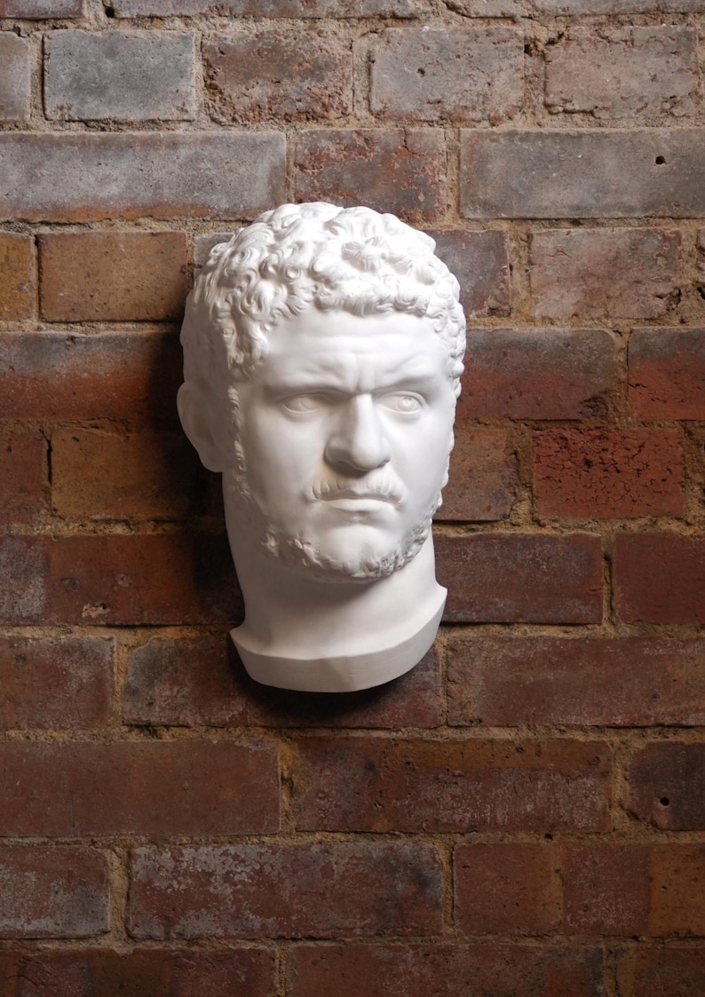 May include: A white plaster bust of a man with curly hair, a mustache, and a beard, mounted on a red brick wall. The sculpture has a stern expression and is a classical art piece.