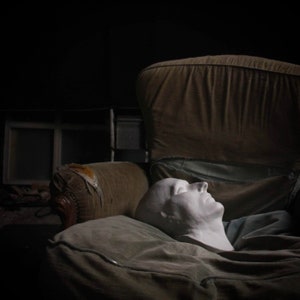 May include: A white plaster cast of a person's face is lying on a brown and beige armchair. The face is closed-eyed and has a relaxed expression.