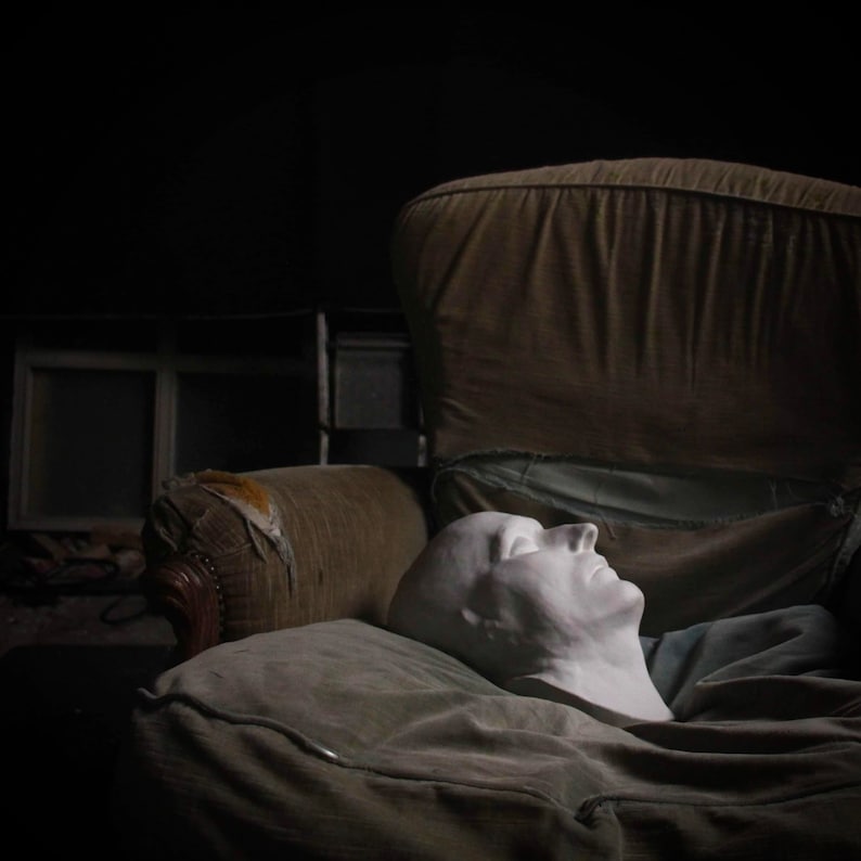 May include: A white plaster cast of a person's face is lying on a brown and beige armchair. The face is closed-eyed and has a relaxed expression.