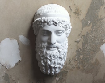 May include: White plaster sculpture of a bearded man's head with a headband. The sculpture is mounted on a wall.