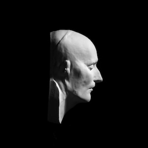 May include: A white plaster cast of a person's head and neck, viewed from the side. The person's eyes are closed and their mouth is slightly open.