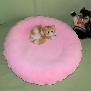 Könnte beinhalten: Ein rundes, weiches, hellrosa Kissen mit einer kleinen, flauschigen, orange-weißen Kätzchenfigur in der Mitte. Rechts neben dem Kissen steht eine schwarz-weiße, katzenförmige Teekanne aus Keramik mit einer rosa Schleife am Hals.