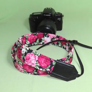 Könnte beinhalten: Eine schwarze Canon EOS 1000F Kamera mit Objektiv, platziert hinter einem aufgerollten Kameragurt. Der Gurt zeigt ein florales Muster mit leuchtend rosa und roten Rosen auf schwarzem Grund und hat schwarze Endstücke in Lederoptik.