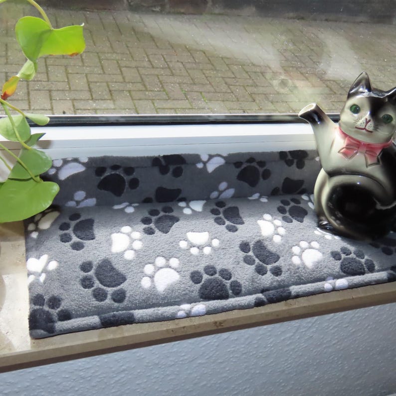 Puede incluir: Un alf&eacute;izar de ventana gris con un coj&iacute;n gris y blanco con estampado de huellas de patas. Una figurita de gato blanco y negro est&aacute; sentada en el coj&iacute;n.