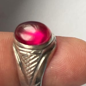 Puede incluir: Un anillo de plata con una gran gema roja brillante de forma ovalada. La banda del anillo tiene un diseño texturizado y detallado. La gema es el punto focal, captando la luz y mostrando un color rico y saturado. El anillo es una pieza de joyería.