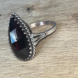 Op de afbeelding: Een zilveren ring met een grote, facetgeslepen, zwarte edelsteen in een druppelvorm. De edelsteen is omgeven door een decoratieve, touwachtige rand. De ringband heeft een eenvoudig, elegant ontwerp.