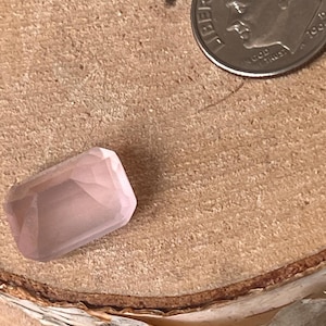 Könnte beinhalten: Ein blassrosa, rechteckiger facettierter Edelstein, der neben einer amerikanischen Zehn-Cent-Münze zum Größenvergleich liegt. Der Edelstein hat eine klare, polierte Oberfläche mit mehreren geometrischen Facetten, die das Licht reflektieren.