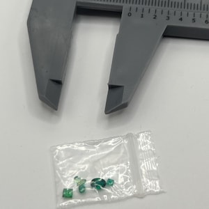 Könnte beinhalten: Ein graues Messschieber-Werkzeug befindet sich über einem kleinen Plastikbeutel mit mehreren kleinen, grünen Edelsteinen. Die Edelsteine haben unterschiedliche Formen, darunter quadratisch, oval und rund, und sind smaragdgrün.