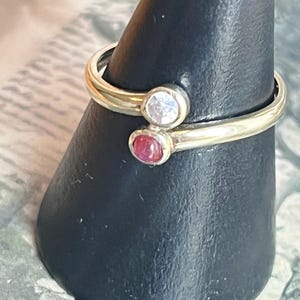 Puede incluir: Dos anillos dorados con piedras preciosas engastadas. Un anillo presenta una piedra preciosa clara y redonda, y el otro tiene una piedra preciosa roja y redonda. Los anillos se exhiben en un soporte negro en forma de cono.