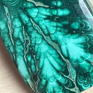 Pode incluir: Close-up de uma pedra de malaquite polida. A pedra apresenta uma cor verde vibrante com padrões intrincados de redemoinhos e faixas verdes claras e escuras. A superfície é lisa e refletora, mostrando a beleza natural do mineral.