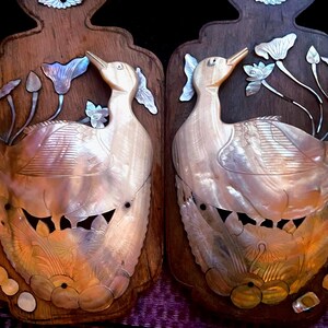 Könnte beinhalten: Zwei dekorative Wandtafeln mit Entenmotiven. Die Enten sind aus schillerndem Material, möglicherweise Perlmutt, gefertigt und vor einem dunklen Holzgrund platziert. Lotusblüten-Details und eine dekorative Oberseite vervollständigen das Design.