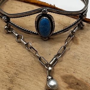 Könnte beinhalten: Silbernes Armreif-Armband mit einem Türkisstein und einer daran hängenden Kette mit einem Perlenanhänger. Das Armband hat aufwendige Details und eine Vintage-Ästhetik. Der Schmuck wird auf einer Holzoberfläche präsentiert.