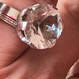 Könnte beinhalten: Ein silberfarbener Ring mit einem großen, runden, klaren facettierten Edelstein in einer Krappenfassung. Die Ringschiene hat eine glatte, polierte Oberfläche und ein zweites, dünnes Banddetail mit kleinen, eingebetteten klaren Steinen an der Seite.