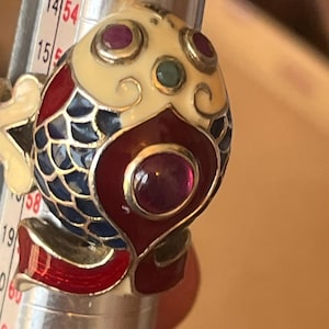 Könnte beinhalten: Ein fischförmiger Ring mit cremefarbenen, roten und blauen Emaille-Schuppen, verziert mit kleinen runden roten und grünen Edelsteinen. Der Ring befindet sich auf einem Ringmaßstab aus Metall und liegt zwischen den Markierungen 15 und 16.