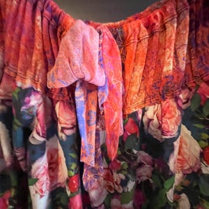 May include: Close-up of a colorful floral top. The top features a vibrant mix of pink, orange, and blue hues. The fabric is adorned with a rose pattern, and a bow detail adds a touch of elegance. The top is made of a lightweight, flowing material.