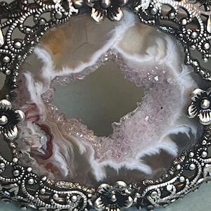 May include: A silver-toned metal cuff bracelet featuring an oval-shaped geode centerpiece with white, gray, and light purple crystalline textures. The frame is decorated with intricate filigree scrollwork and small metal flower accents.