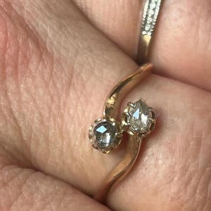 Könnte beinhalten: Ein Bypass-Ring aus rosafarbenem Metall mit zwei facettierten Steinen in Krappenfassung. Die Ringschiene schwingt sich um den Finger und positioniert die Steine nebeneinander. Ein separater, dünner, silberfarbener Ring mit kleinen Steinen ist sichtbar.