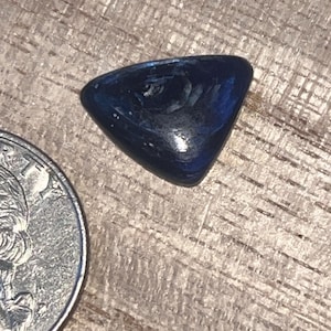 May include: A polished, triangular-shaped dark blue gemstone with a smooth, reflective surface. The stone is positioned next to a United States quarter coin for scale, showing its relative size in comparison to the standard currency.