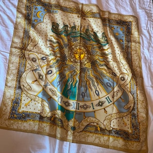 May include: A square silk scarf featuring a central golden sun face surrounded by rays, Roman numerals, and celestial motifs including stars and a crescent moon. The design incorporates blue, gold, and cream tones with the text HERMES PARIS.
