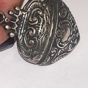 Peut inclure: Gros plan sur un pendentif argenté en forme de cœur avec des motifs tourbillonnants complexes. Le pendentif présente un détail en forme de couronne en haut et une petite boucle pour une chaîne. Le design est détaillé et orné, de style vintage ou antique.