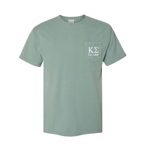 Puede incluir: Camiseta verde salvia de manga corta con un bolsillo en el pecho izquierdo. El bolsillo presenta las letras blancas "K&Sigma;" sobre el texto "Est. 1869". La camiseta est&aacute; hecha de un material suave y tiene un cuello redondo cl&aacute;sico.