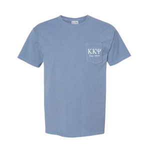 May include: A light blue t-shirt with a pocket on the left side. The pocket has white text that reads "KK&Psi; Est. 1919".