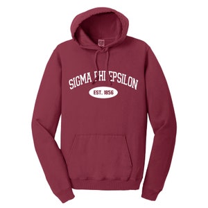 May include: A maroon hooded sweatshirt with a front pocket and drawstring. The white text "SIGMA PHI EPSILON" arches above an oval that reads "EST. 1856". The sweatshirt has a hood and is made of a soft material.