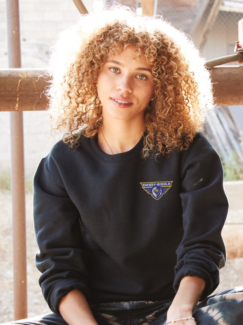 Pode incluir: Uma mulher vestindo uma camisa de moletom preta com um logotipo bordado em azul e amarelo que diz "Embry-Riddle".
