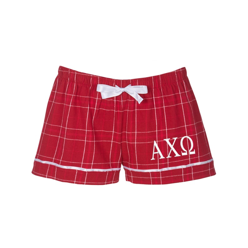 Puede incluir: Pantalones de pijama de cuadros rojos con un lazo blanco y letras bordadas blancas "AXO" en la pierna derecha.