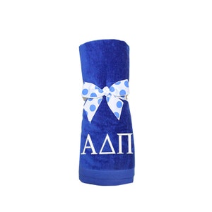 May include: A royal blue beach towel with a white bow tied around it. The bow has blue polka dots. The towel has the letters "ΑΔΠ" embroidered on it in white.