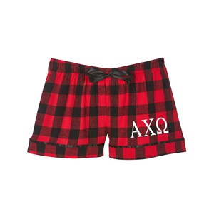 Puede incluir: Pantalones de pijama de cuadros rojos y negros con un lazo negro y letras bordadas blancas que dicen "AXO".