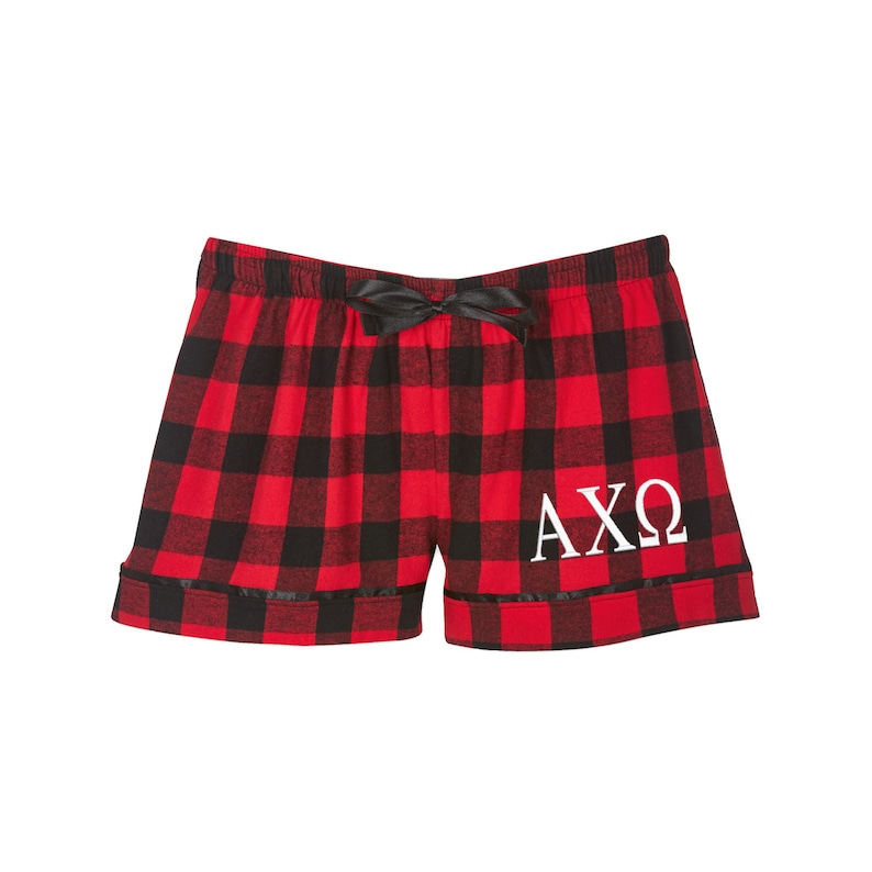 Puede incluir: Pantalones de pijama de cuadros rojos y negros con un lazo negro y letras bordadas blancas que dicen "AXO".