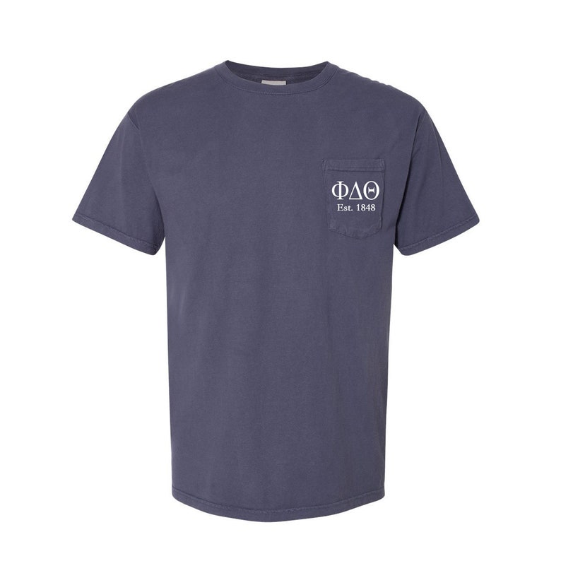 May include: A dark blue t-shirt with a pocket on the left side. The pocket has white embroidery that reads "&Phi;&Delta;&Theta; Est. 1848".