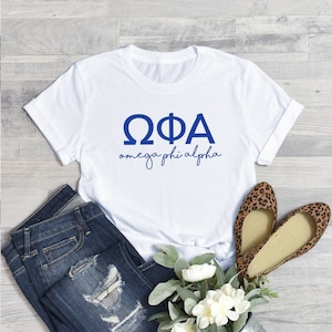 May include: White t-shirt with blue lettering that reads "ΩΦΑ omega phi alpha". The shirt is shown on a wooden surface with a pair of jeans and a bouquet of white flowers.