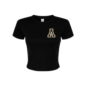 May include: Black cropped t-shirt with a yellow embroidered letter "A" on the left chest. The letter has a white outline and the word "Atlanta" is written inside the letter.