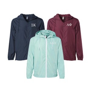 May include: Three hooded windbreakers in different colors: navy blue, burgundy, and light blue. Each jacket has a white zipper and a white embroidered Greek letter design on the front. The navy blue jacket has "ΣΚ" embroidered on the front, the burgundy jacket has "ΑΦ" embroidered on the front, and the light blue jacket has "ΑΠΠ" embroidered on the front.