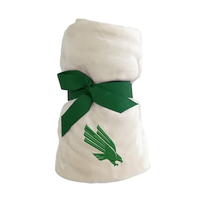 May include: A rolled-up, cream-colored plush blanket tied with a wide, dark green ribbon. The blanket features an embroidered green eagle logo, adding a touch of collegiate spirit. The soft texture and vibrant accents make it a cozy gift.