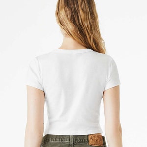 K&ouml;nnte beinhalten: Wei&szlig;es, bauchfreies T-Shirt mit kurzen &Auml;rmeln. Das T-Shirt wird von einer Person mit langen blonden Haaren getragen. Die Person tr&auml;gt eine gr&uuml;ne Jeans mit einem braunen Lederflicken auf der Ges&auml;&szlig;tasche, auf dem "Gap Denim" steht.