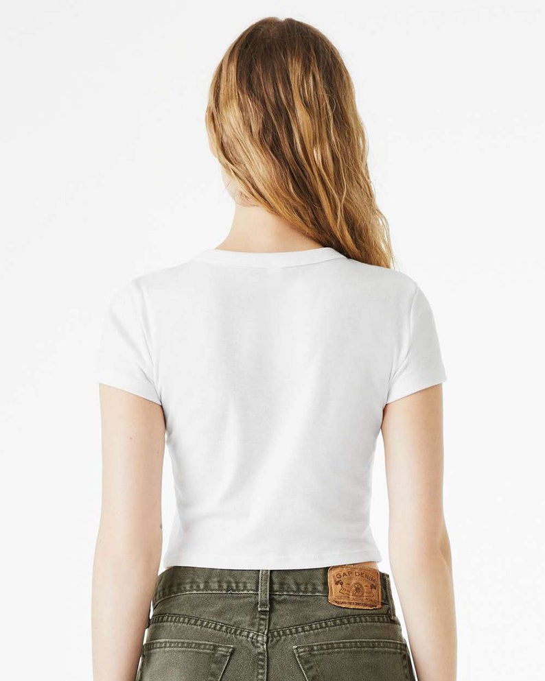 K&ouml;nnte beinhalten: Wei&szlig;es, bauchfreies T-Shirt mit kurzen &Auml;rmeln. Das T-Shirt wird von einer Person mit langen blonden Haaren getragen. Die Person tr&auml;gt eine gr&uuml;ne Jeans mit einem braunen Lederflicken auf der Ges&auml;&szlig;tasche, auf dem "Gap Denim" steht.